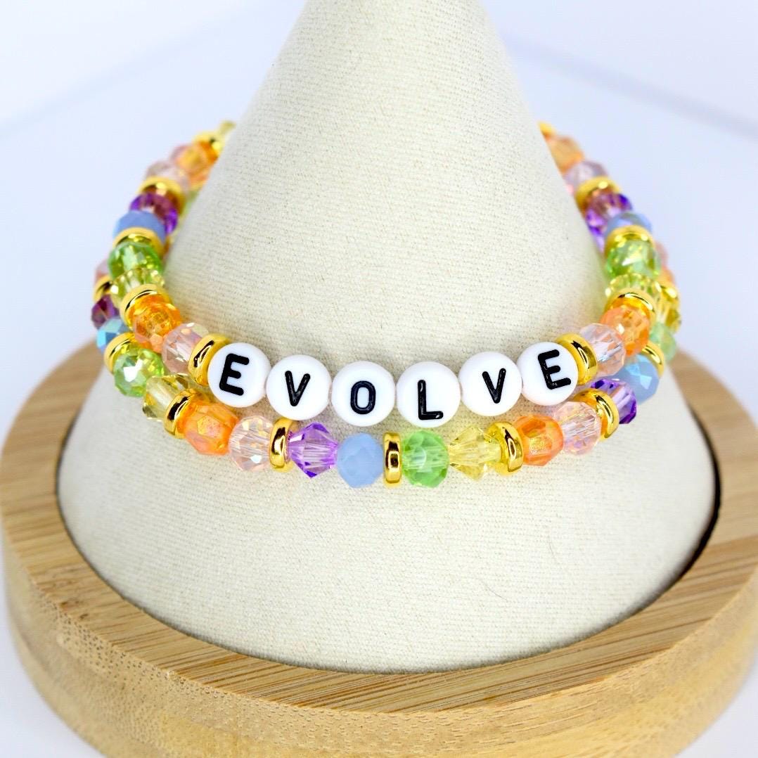 Two beaded bracelets with the word "EVOLVE" spelled out in white letter beads. The bracelets feature colorful beads in shades of orange, yellow, green, purple, and blue, with gold accents. The bracelets are displayed on a white cone-shaped stand.