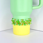 A light green tumbler with a yellow base, adorned with three beaded bracelets. The bracelets feature green beads and gold accents, including heart-shaped charms. The tumbler has a handle and is set against a white background.