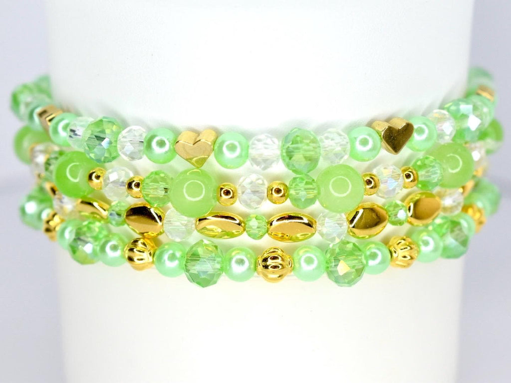 A stack of beaded bracelets in shades of green, clear, and gold. The bracelets feature various bead shapes, including round, faceted, and heart-shaped beads. The gold beads add a touch of elegance to the design.