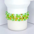 Four beaded bracelets in shades of green and clear, with gold-colored accents. The bracelets are made of various sizes and shapes of beads, including round, oval, and heart-shaped beads. The bracelets are stacked on a white cylindrical object.