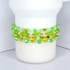 Four beaded bracelets in shades of green and clear, with gold-colored accents. The bracelets are made of various sizes and shapes of beads, including round, oval, and heart-shaped beads. The bracelets are stacked on a white cylindrical object.