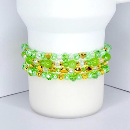 Four beaded bracelets in shades of green and clear, with gold-colored accents. The bracelets are made of various sizes and shapes of beads, including round, oval, and heart-shaped beads. The bracelets are stacked on a white cylindrical object.