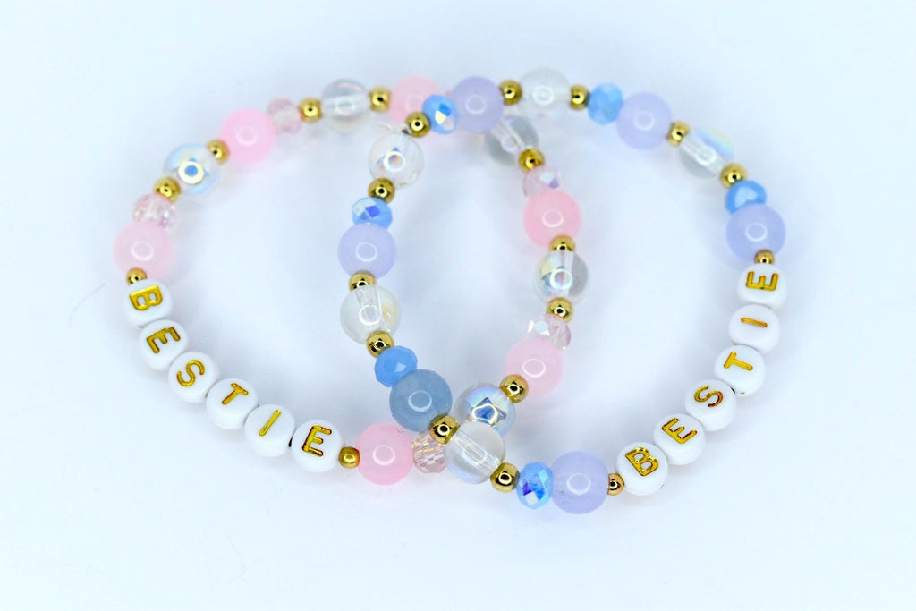 Two beaded bracelets with the word 'BESTIE' spelled out in gold letters on white beads. The bracelets feature a mix of pink, blue, and clear beads with gold accents. The beads are arranged in a circular pattern, creating a friendship bracelet design.