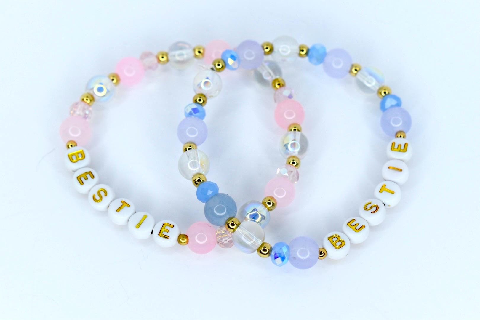 Two beaded bracelets with the word 'BESTIE' spelled out in gold letters on white beads. The bracelets feature a mix of pink, blue, and clear beads with gold accents. The beads are arranged in a circular pattern, creating a friendship bracelet design.