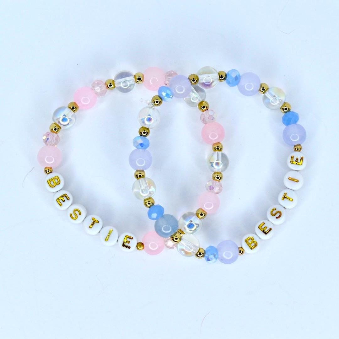 Two beaded bracelets with the word 'BESTIE' spelled out in white letter beads. The bracelets feature a mix of pink, blue, and clear beads with gold accents. The beads are arranged in a pattern, creating a colorful and stylish accessory. The bracelets are likely for friendship or as a gift.