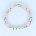 A pink beaded bracelet with the word 'BESTIE' spelled out in white letter beads with gold lettering. The bracelet features a mix of pink, clear, and iridescent beads, along with gold accents.