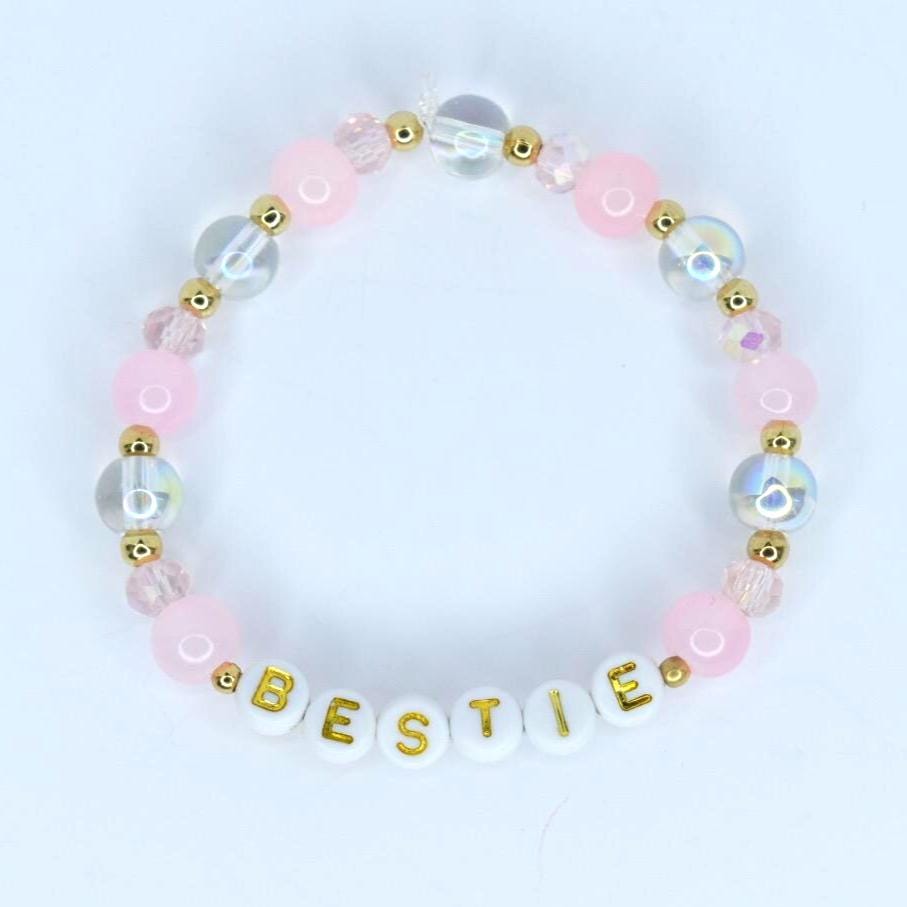 A pink beaded bracelet with the word 'BESTIE' spelled out in white letter beads with gold lettering. The bracelet features a mix of pink, clear, and iridescent beads, along with gold accents.