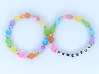 Two beaded bracelets with a rainbow of colors. One bracelet spells out the word "POWERFUL" in white letter beads. The bracelets feature a variety of colorful beads, including pink, orange, yellow, green, blue, and purple.
