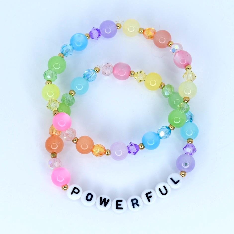 Two colorful beaded bracelets. The bracelets feature a variety of pastel-colored beads, including pink, orange, yellow, green, blue, and purple. One bracelet has the word "POWERFUL" spelled out in white letter beads. The bracelets are connected with gold-colored accents.