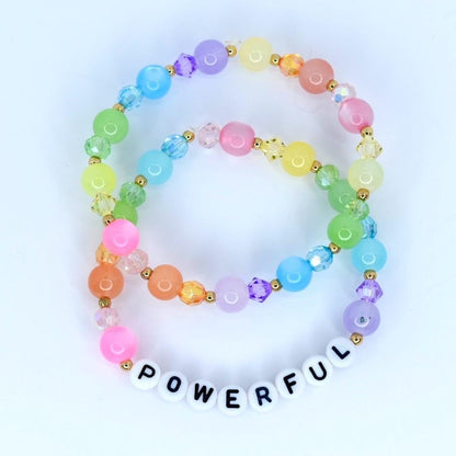 Two colorful beaded bracelets. The bracelets feature a variety of pastel-colored beads, including pink, orange, yellow, green, blue, and purple. One bracelet has the word "POWERFUL" spelled out in white letter beads. The bracelets are connected with gold-colored accents.