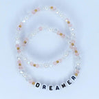 Two beaded bracelets. One bracelet is made of clear and pink beads with gold accents. The second bracelet spells out the word "DREAMER" with white letter beads.