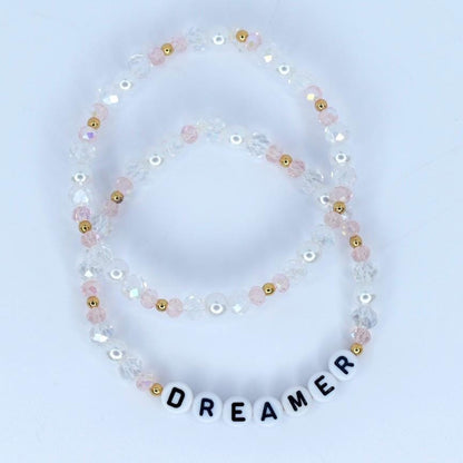 Two beaded bracelets. One bracelet is made of clear and pink beads with gold accents. The second bracelet spells out the word "DREAMER" with white letter beads.