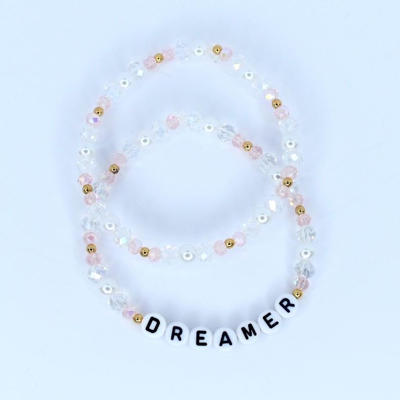 Two beaded bracelets. The bracelets are made of clear, pink, and gold beads. One bracelet has the word "DREAMER" spelled out in white letter beads. The bracelets are on a white background.