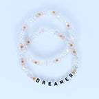 Two beaded bracelets. The bracelets are made of clear, pink, and gold beads. One bracelet has the word "DREAMER" spelled out in white letter beads. The bracelets are on a white background.