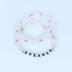 Two beaded bracelets. One bracelet is clear with pink and gold beads. The other bracelet is clear with pink, gold, and white beads. The bottom bracelet has white letter beads that spell out the word "DREAMER".