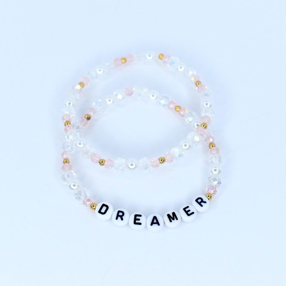 Two beaded bracelets. One bracelet is clear with pink and gold beads. The other bracelet is clear with pink, gold, and white beads. The bottom bracelet has white letter beads that spell out the word "DREAMER".