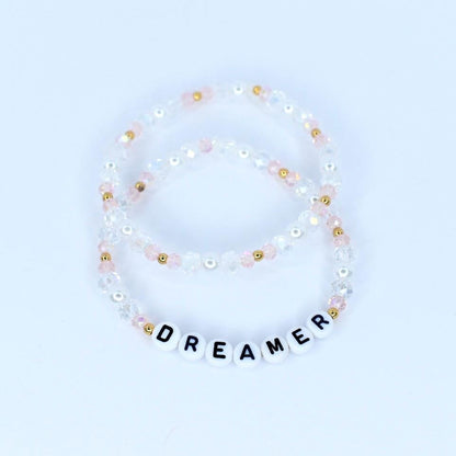 Two beaded bracelets. One bracelet is clear with pink and gold beads. The other bracelet is clear with pink, gold, and white beads. The bottom bracelet has white letter beads that spell out the word "DREAMER".