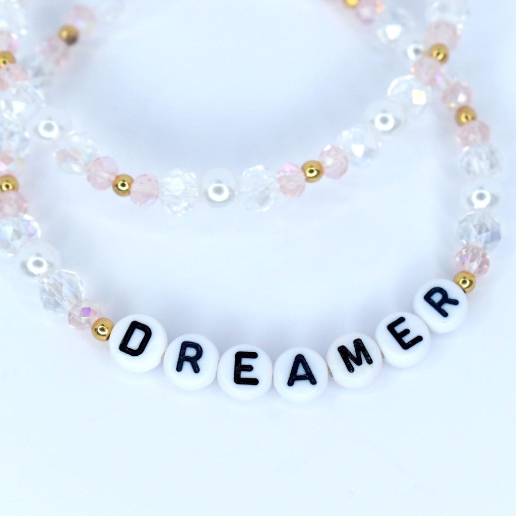 Two beaded bracelets. The top bracelet features clear and pink beads with gold accents. The bottom bracelet spells out the word "DREAMER" with white letter beads. The bracelets are on a white background.