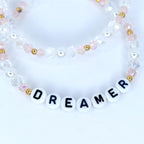 Two beaded bracelets. The top bracelet features clear and pink beads with gold accents. The bottom bracelet spells out the word "DREAMER" with white letter beads. The bracelets are on a white background.