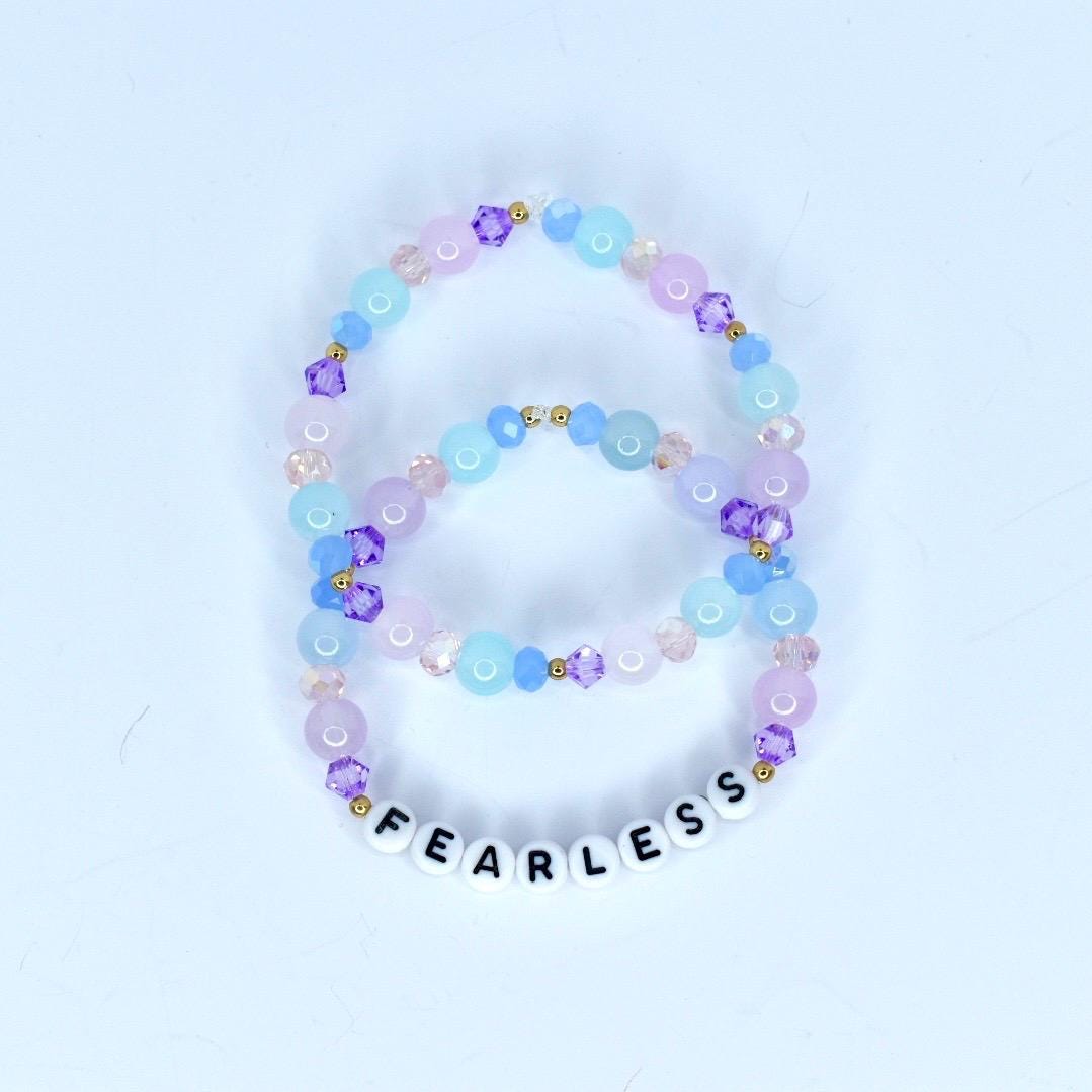 Two beaded bracelets in shades of purple, pink, and blue. The bracelets feature a mix of round and faceted beads, with the word 'FEARLESS' spelled out in white letter beads on the bottom bracelet. The beads are strung on a clear elastic cord.