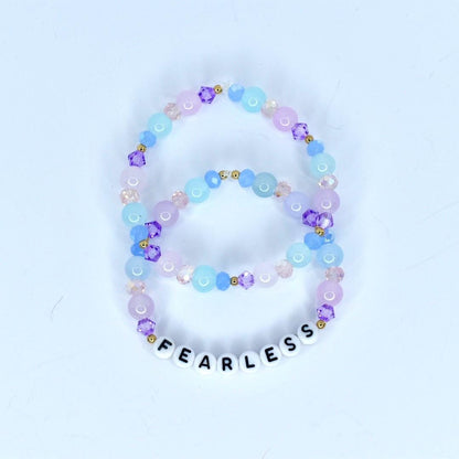 Two beaded bracelets in shades of purple, pink, and blue. The bracelets feature a mix of round and faceted beads, with the word 'FEARLESS' spelled out in white letter beads on the bottom bracelet. The beads are strung on a clear elastic cord.