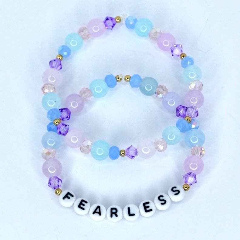 Two beaded bracelets in shades of blue, purple, and pink. The bracelets feature round and faceted beads, with the bottom bracelet spelling 'FEARLESS' in white letter beads. Gold-colored accents are interspersed throughout the design.