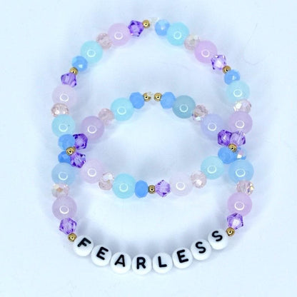 Two beaded bracelets in shades of blue, purple, and pink. The bracelets feature round and faceted beads, with the bottom bracelet spelling 'FEARLESS' in white letter beads. Gold-colored accents are interspersed throughout the design.