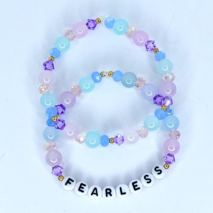 Two beaded bracelets in shades of blue, purple, and pink. The bracelets feature round and faceted beads, with gold accents. The bottom bracelet spells 'FEARLESS' in white letter beads. The bracelets are likely fashion accessories.
