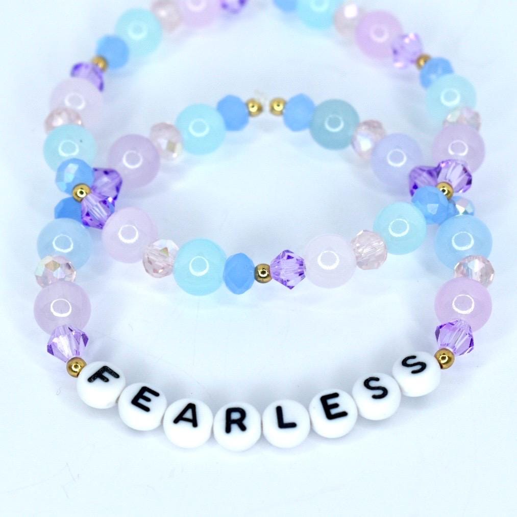 Two beaded bracelets in shades of blue, purple, and pink. The bracelets are adorned with round and faceted beads, and gold accents. One bracelet features white letter beads spelling 'FEARLESS'.