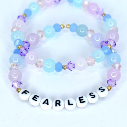 Two beaded bracelets in shades of blue, purple, and pink. The bracelets are adorned with round and faceted beads, and gold accents. One bracelet features white letter beads spelling 'FEARLESS'.