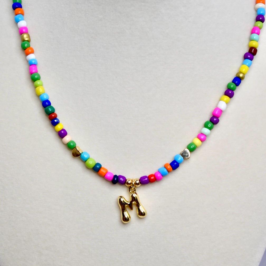 Close-up of colorful beaded necklace with gold letter M pendant on neutral background.