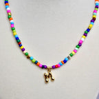 Close-up of colorful beaded necklace with gold letter M pendant on neutral background.