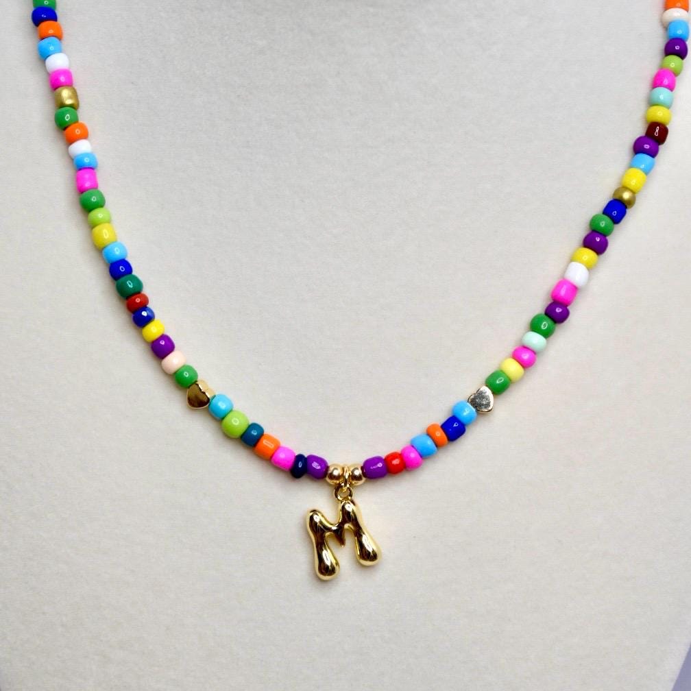 Close-up of colorful beaded necklace with gold letter M pendant on neutral background.
