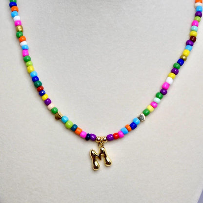 Close-up of colorful beaded necklace with gold letter M pendant on neutral background.