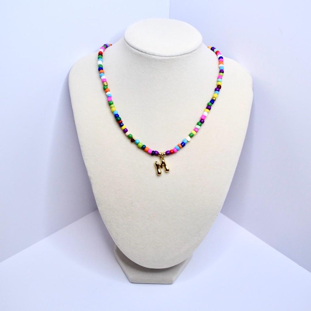 Colorful beaded necklace with gold letter M charm displayed on white velvet jewelry stand.