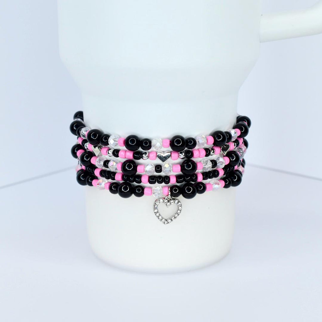 A stack of beaded bracelets in black, pink, and clear beads. Each bracelet has a different pattern, with silver heart charms dangling from the bottom. The bracelets are stacked on a white cylindrical object.