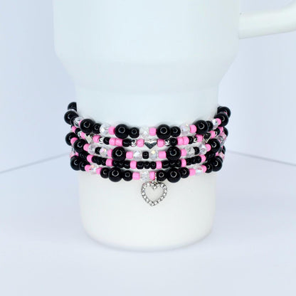 A stack of beaded bracelets in black, pink, and clear beads. Each bracelet has a different pattern, with silver heart charms dangling from the bottom. The bracelets are stacked on a white cylindrical object.