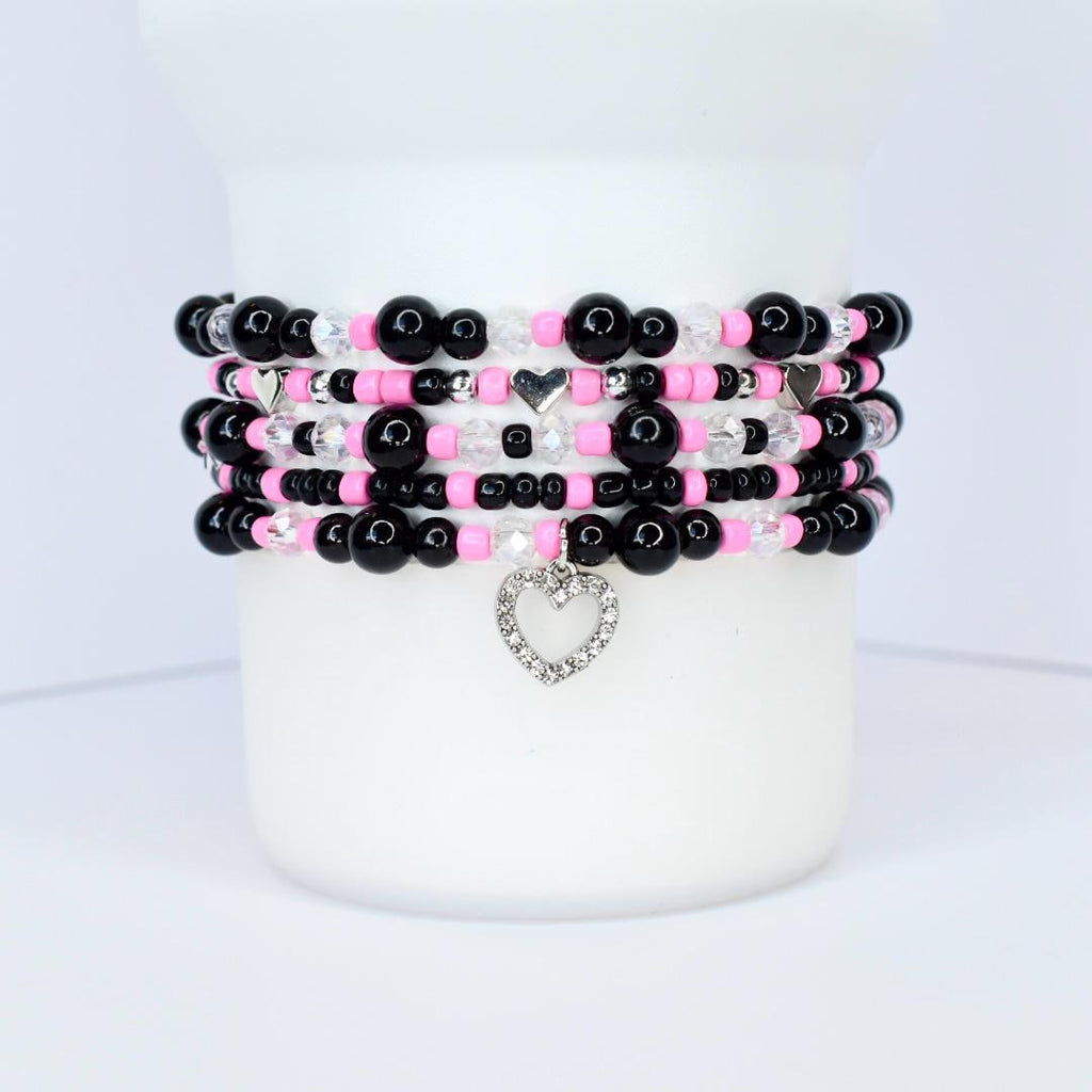 Stack of beaded bracelets in black, pink, and clear beads. Each bracelet has a different design, with some featuring silver heart charms. The bracelets are stacked on a white cylindrical surface.