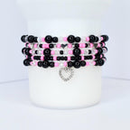 Stack of beaded bracelets in black, pink, and clear beads. Each bracelet has a different design, with some featuring silver heart charms. The bracelets are stacked on a white cylindrical surface.