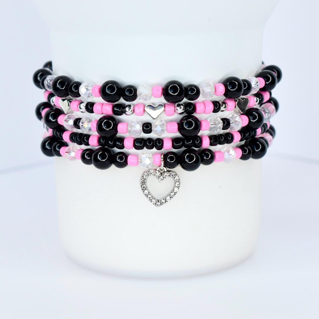 Stack of beaded bracelets in black, pink, and clear beads. Each bracelet has a different design, with silver heart charms and accents. The bracelets are stacked on a white surface.