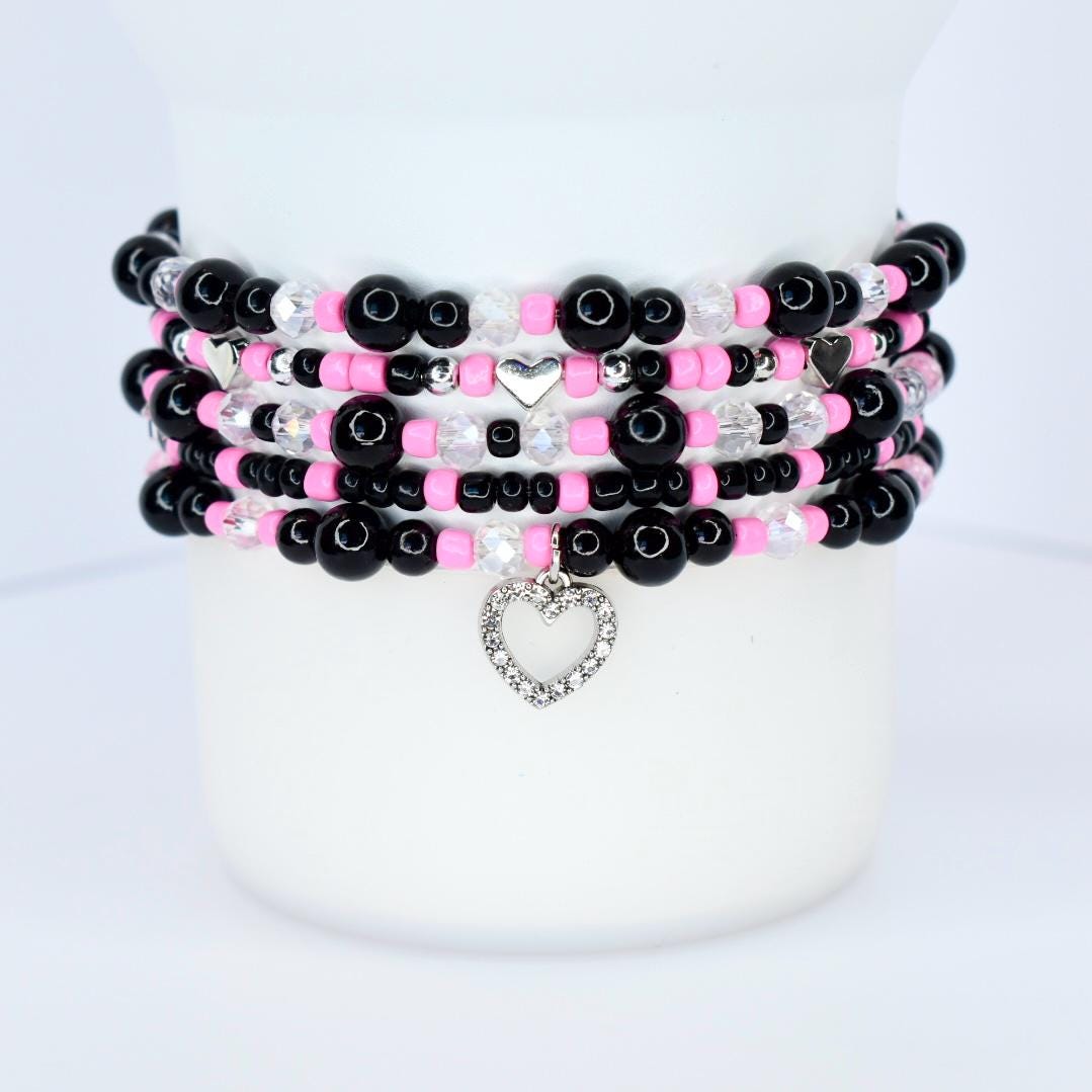 Stack of beaded bracelets in black, pink, and clear beads. Each bracelet has a different design, with silver heart charms and accents. The bracelets are stacked on a white surface.