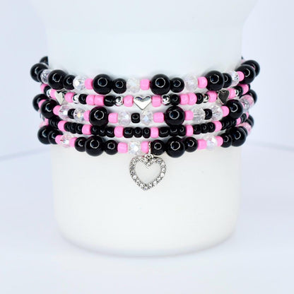 Stack of beaded bracelets in black, pink, and clear beads. Each bracelet has a different design, with silver heart charms and accents. The bracelets are stacked on a white surface.