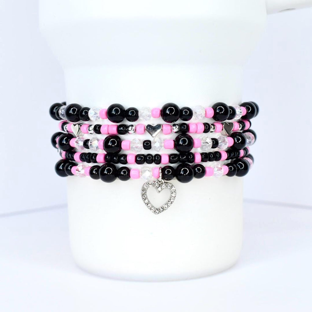 Stack of beaded bracelets in black, pink, and clear beads. Each bracelet features a mix of round and faceted beads, with silver heart charms. The bracelets are displayed on a white cylindrical object.