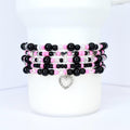 Stack of beaded bracelets in black, pink, and clear beads. Each bracelet features a mix of round and faceted beads, with silver heart charms. The bracelets are displayed on a white cylindrical object.