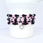 Stack of beaded bracelets in black, pink, and clear beads. Each bracelet features a mix of round and faceted beads, with silver heart charms. The bracelets are displayed on a white cylindrical object.