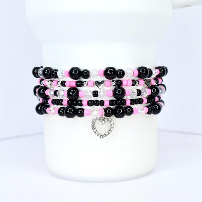 Stack of beaded bracelets in black, pink, and clear beads. Each bracelet features a mix of round and faceted beads, with silver heart charms. The bracelets are displayed on a white cylindrical object.
