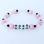 A pink and white beaded bracelet. The bracelet features pink, round beads, clear crystal-like beads, and small black beads. The word 'BOSS' is spelled out with white square beads. The bracelet is a fashionable accessory.