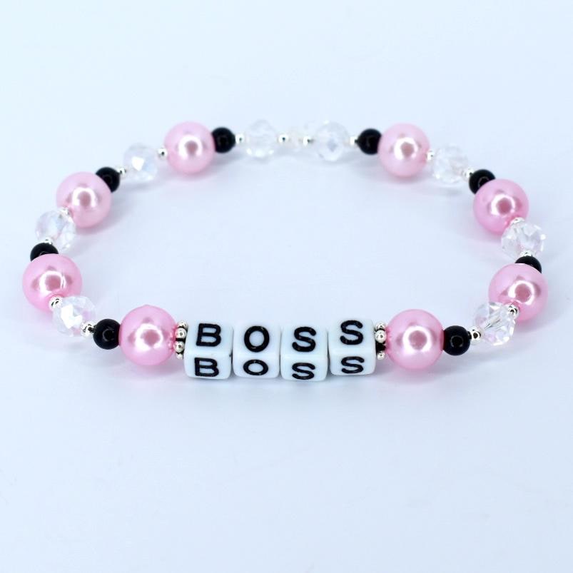 A pink and white beaded bracelet. The bracelet features pink, round beads, clear crystal-like beads, and small black beads. The word 'BOSS' is spelled out with white square beads. The bracelet is a fashionable accessory.