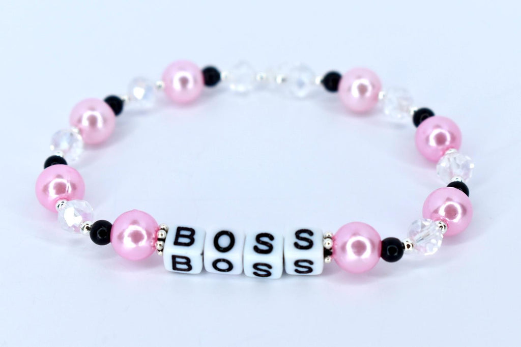 A pink and white beaded bracelet with the word 'BOSS' spelled out in white letter beads. The bracelet features pink pearl beads, clear crystal beads, and black accent beads. The beads are strung together on a stretchy cord, forming a circular shape.