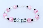 A pink and white beaded bracelet with the word 'BOSS' spelled out in white letter beads. The bracelet features pink pearl beads, clear crystal beads, and black accent beads. The beads are strung together on a stretchy cord, forming a circular shape.